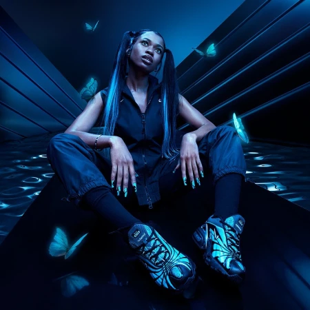 A Black female model with long hair, stylish nail art, and athletic shoes poses in a neon blue studio with glowing butterfly accents.