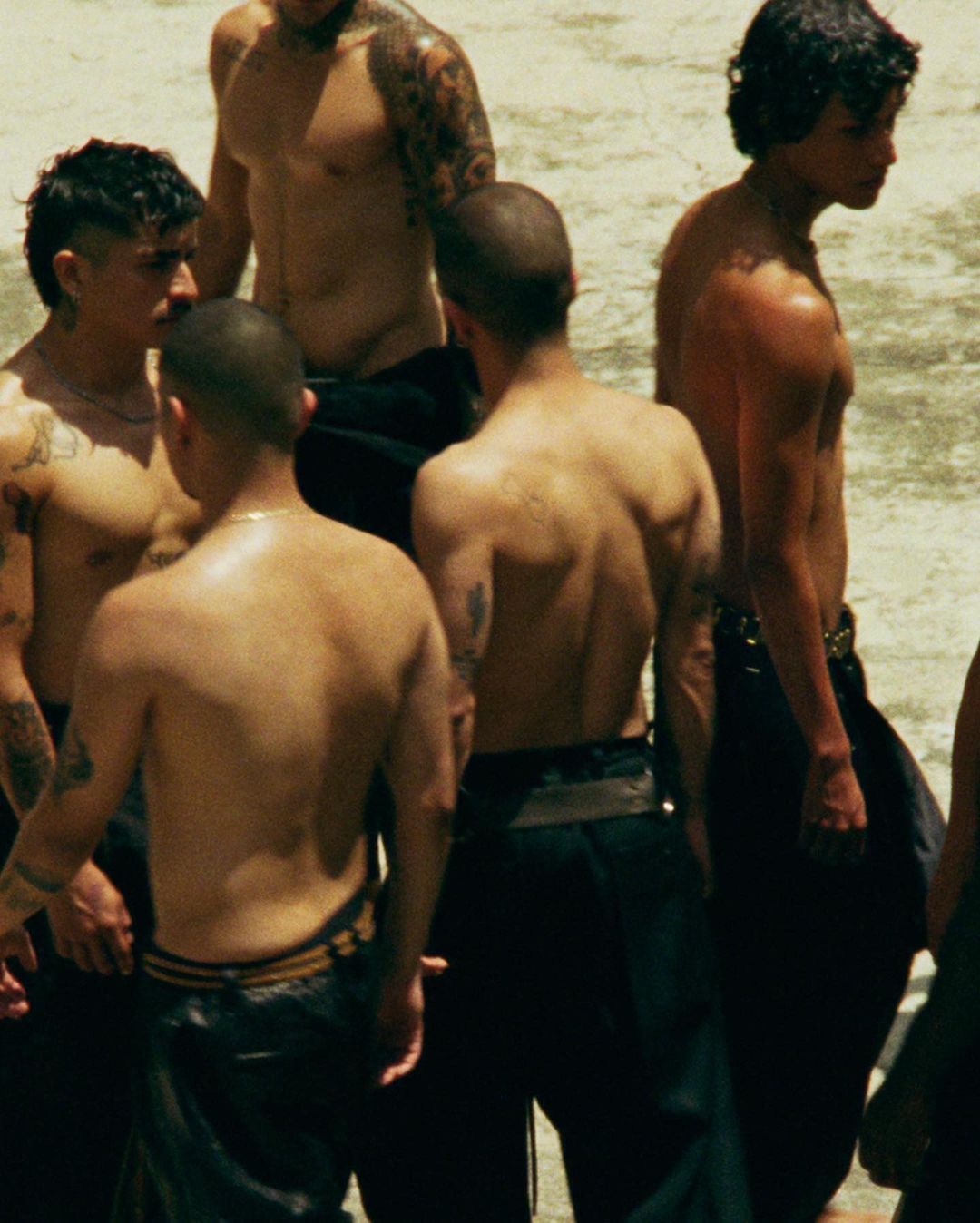 A group of shirtless adult men stand in a semi-circle in an outdoor setting, showing diverse hairstyles, tattoos, and black pants.