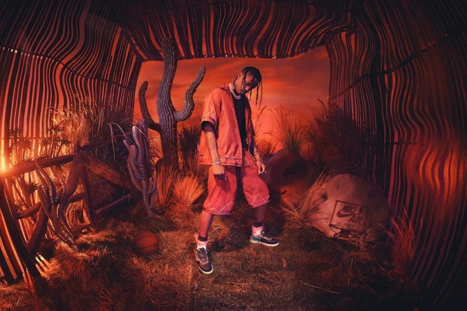 Man in red outfit poses in a surreal desert setting inside, framed by wood, with cacti, grasses, basketball, and Nike shoe box.