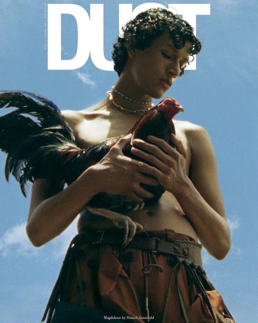 Shirtless person holds a rooster against a blue sky, magazine cover. Leather skirt and a thick necklace adorn the figure.