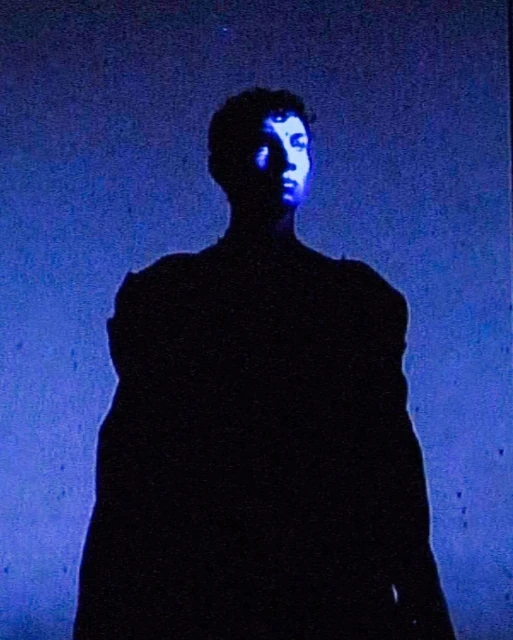 A silhouetted young adult is partially lit with a blue backlight in a dimly lit, low-angle composition.