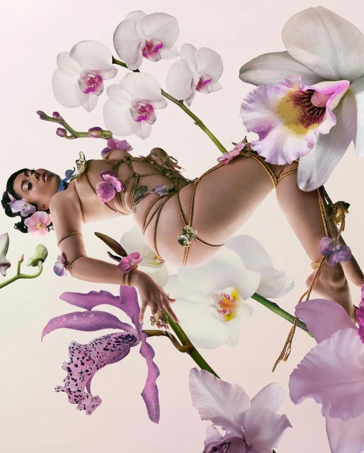 Woman in an ornate gold body chain lying among purple and white orchid flowers in an eye-level studio portrait on a gradient backdrop.