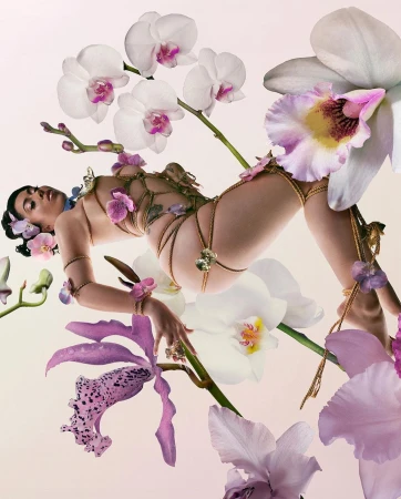 Woman in an ornate gold body chain lying among purple and white orchid flowers in an eye-level studio portrait on a gradient backdrop.