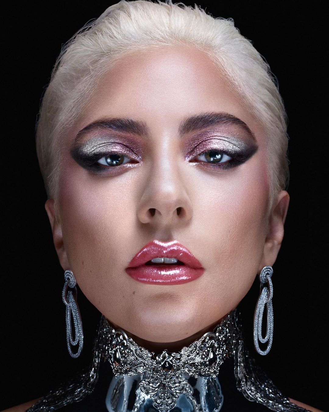 Close-up portrait of a fair-skinned woman with short platinum blonde hair, elaborate silver eye makeup, and statement jewelry against black.