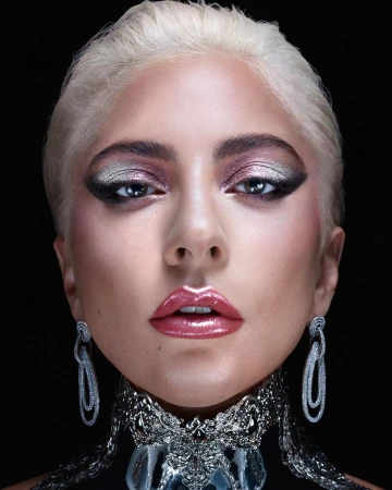 Close-up portrait of a fair-skinned woman with short platinum blonde hair, elaborate silver eye makeup, and statement jewelry against black.