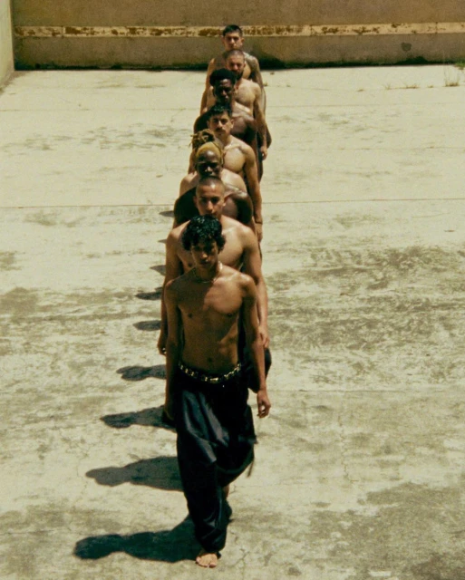 Group of shirtless men standing in a line outdoors, one wearing loose black pants, captured in a medium shot with an eye-level angle.