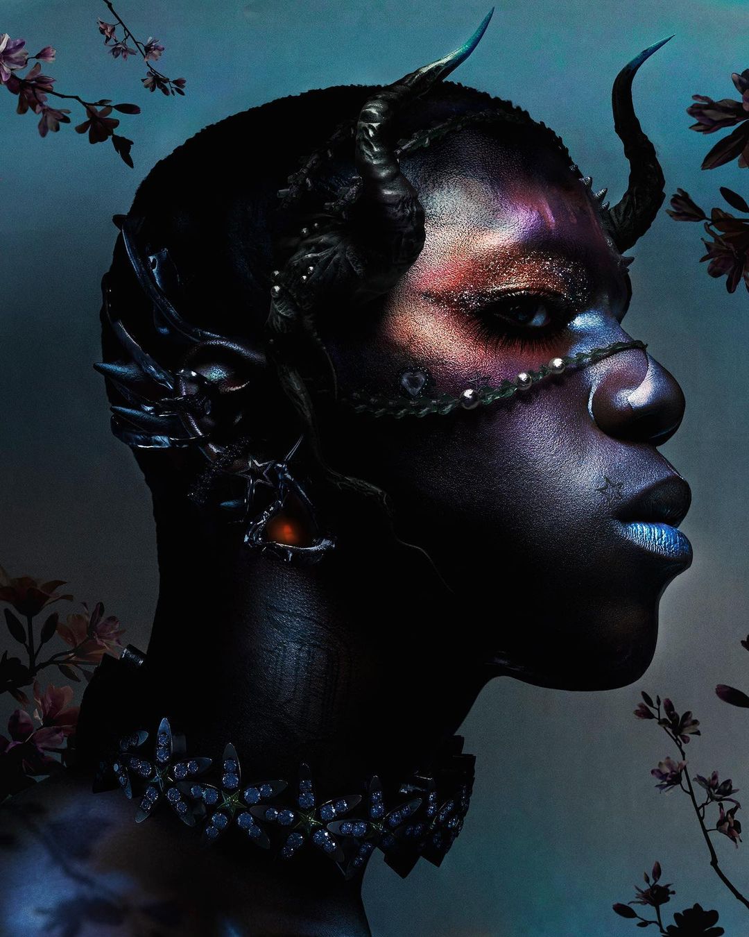 A profile portrait showcases a person with dark skin, adorned with dramatic makeup, horn-like headwear, and an elaborate jeweled collar.