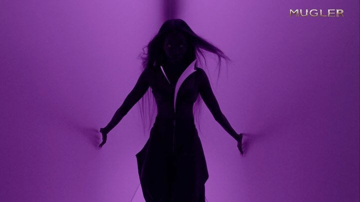 A dark-skinned woman with long hair stands in front of a purple wall with the Mugler logo in the upper right corner.