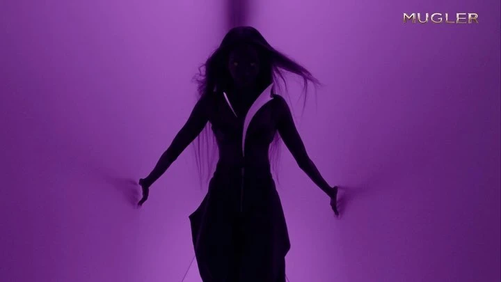 A dark-skinned woman with long hair stands in front of a purple wall with the Mugler logo in the upper right corner.