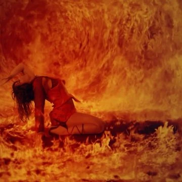 Adult female dancer in red costume lies on fire-covered ground with arms raised and long dark hair flowing in a surreal, artistic shot.