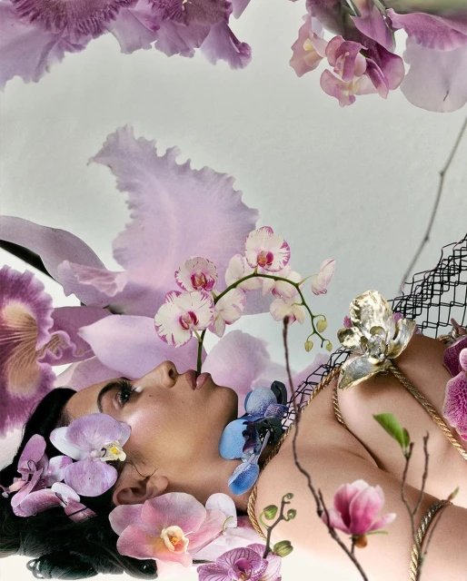 Woman lies among pastel purple, pink, and white orchids. She has dark hair and fair skin and wears a gold necklace and black net headpiece.