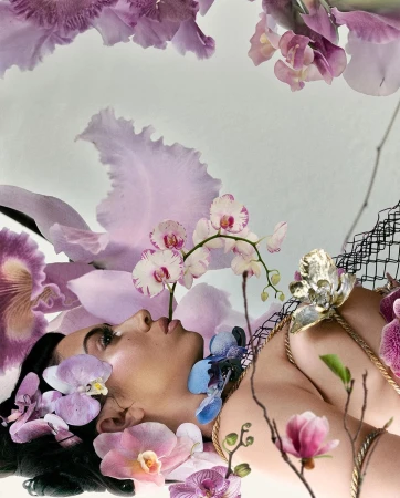 Woman lies among pastel purple, pink, and white orchids. She has dark hair and fair skin and wears a gold necklace and black net headpiece.