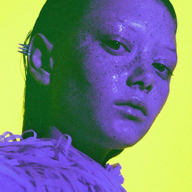 Portrait of a person in purple and yellow light with glittery skin, an ear cuff and lace details on the shoulder in a close-up shot.