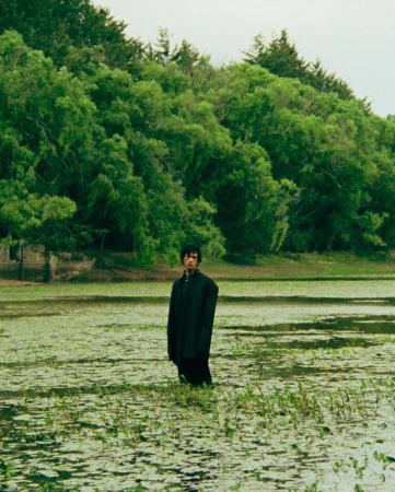 A person wearing a dark coat standing in a shallow body of water surrounded by lush green trees.