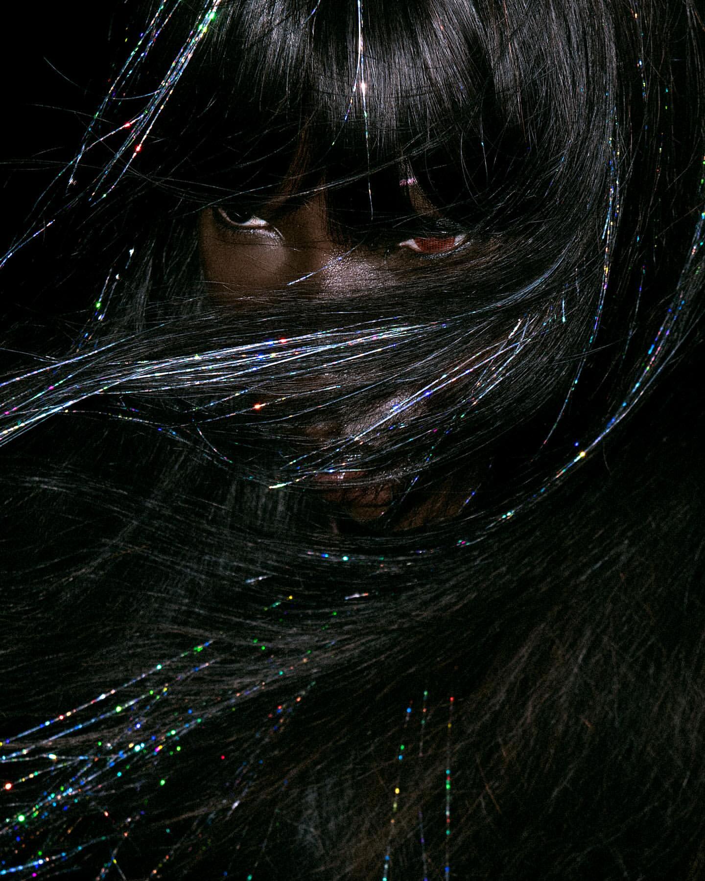 Close-up of a Black woman with dark hair partially covering her face, with shiny tinsel threads woven throughout, against a dark background.