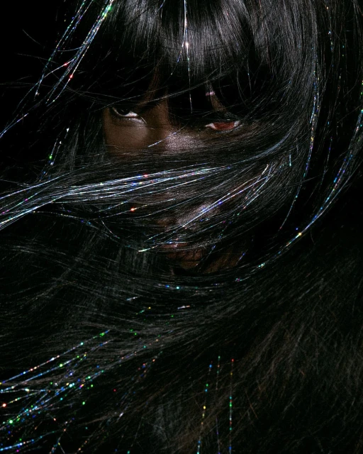 Close-up of a Black woman with dark hair partially covering her face, with shiny tinsel threads woven throughout, against a dark background.