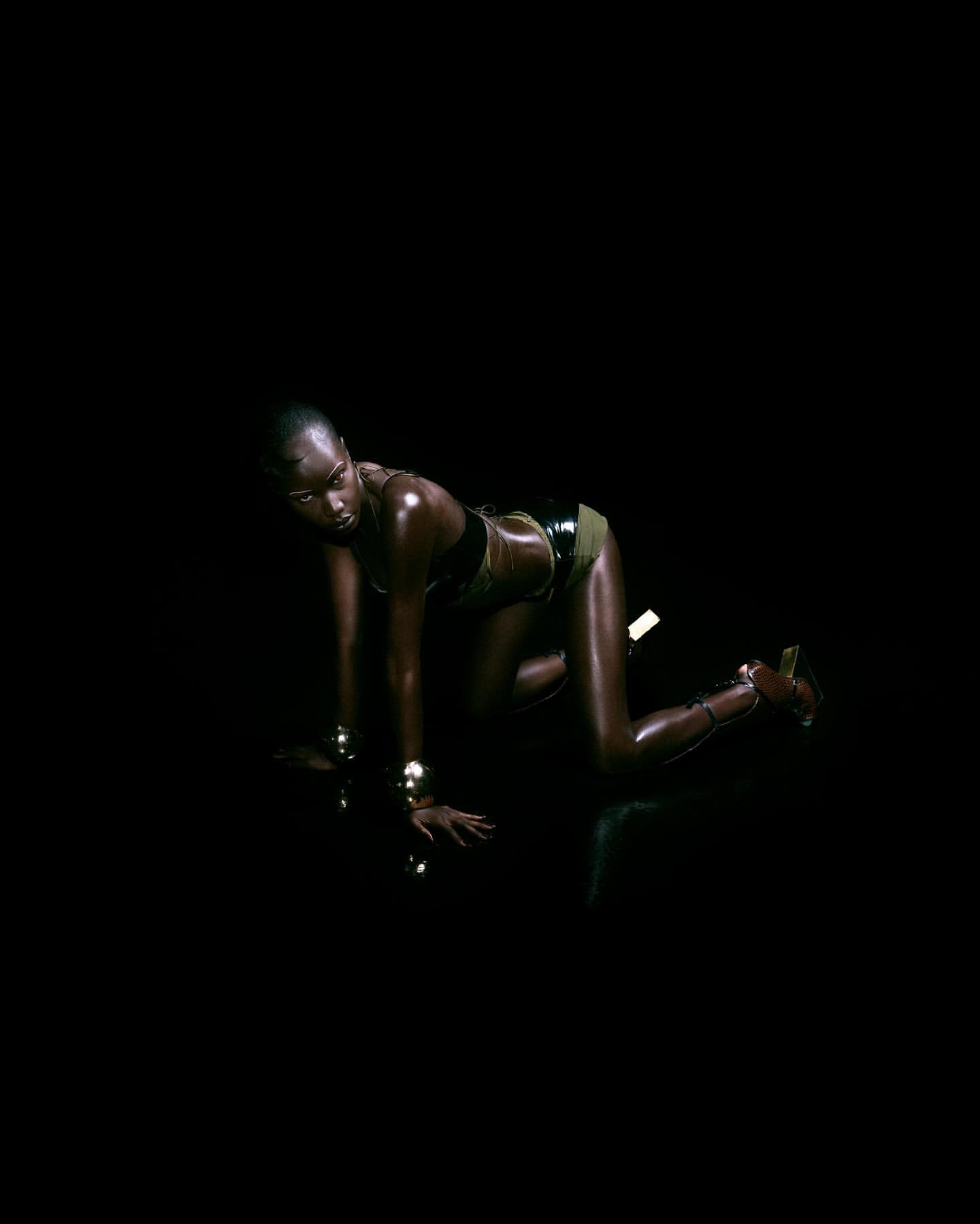 A dark-skinned woman with a shaved head poses in a black two-piece outfit with gold bracelets in a dark studio setting.