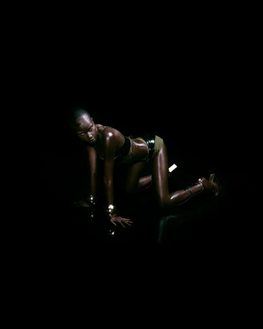 A dark-skinned woman with a shaved head poses in a black two-piece outfit with gold bracelets in a dark studio setting.