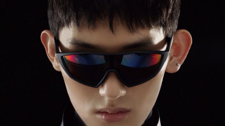 Close-up studio shot of adult male with short black hair wearing black futuristic sunglasses against a dark black background.