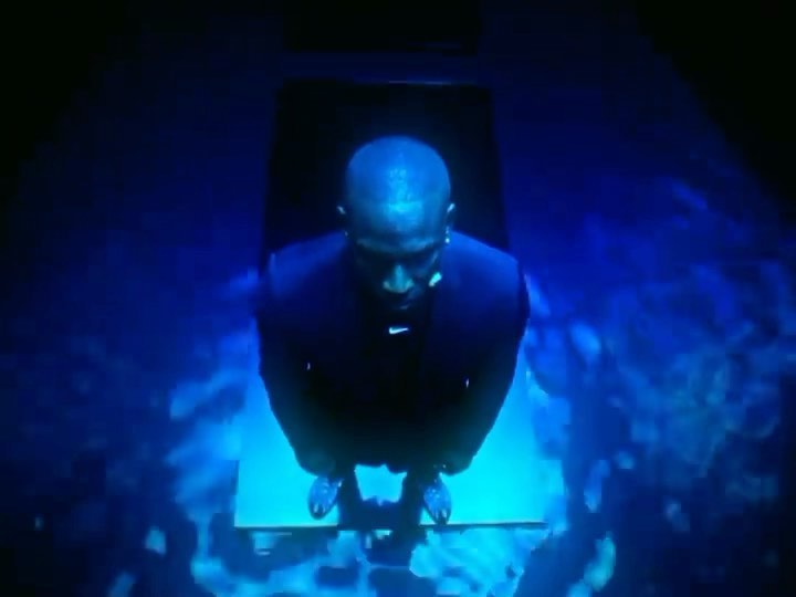 Man in dark suit stands on a light panel in dark room, looking up with dim blue lighting illuminating him from beneath.