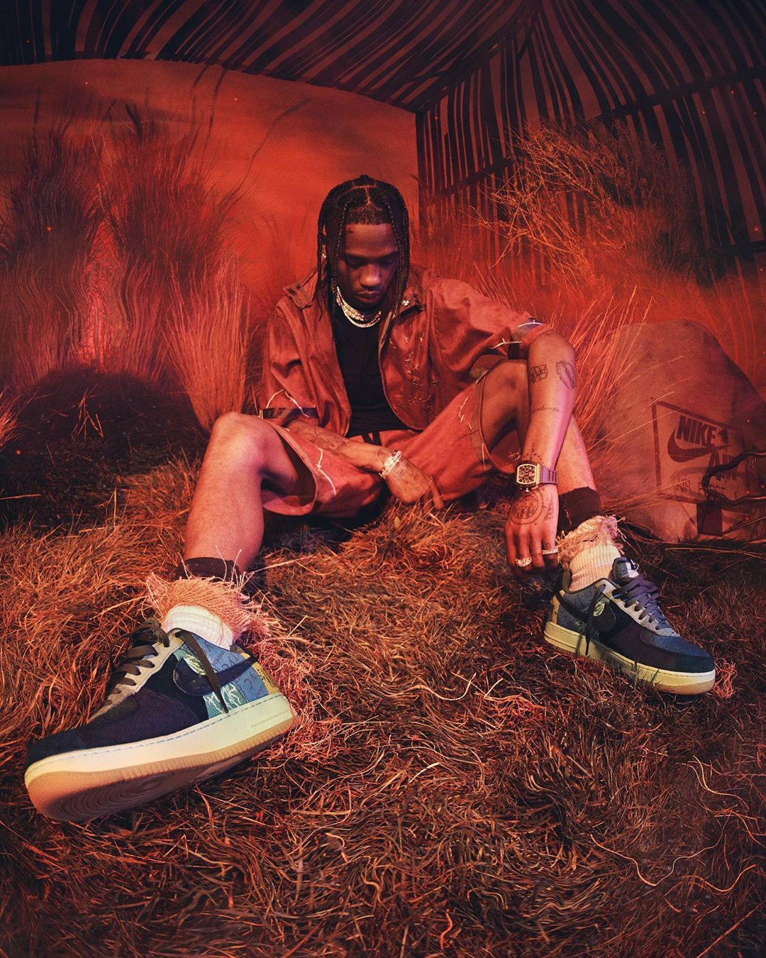 Travis Scott poses in a coral jacket, black tee, and red shorts on a straw covered surface with two Nike sneakers at the forefront.