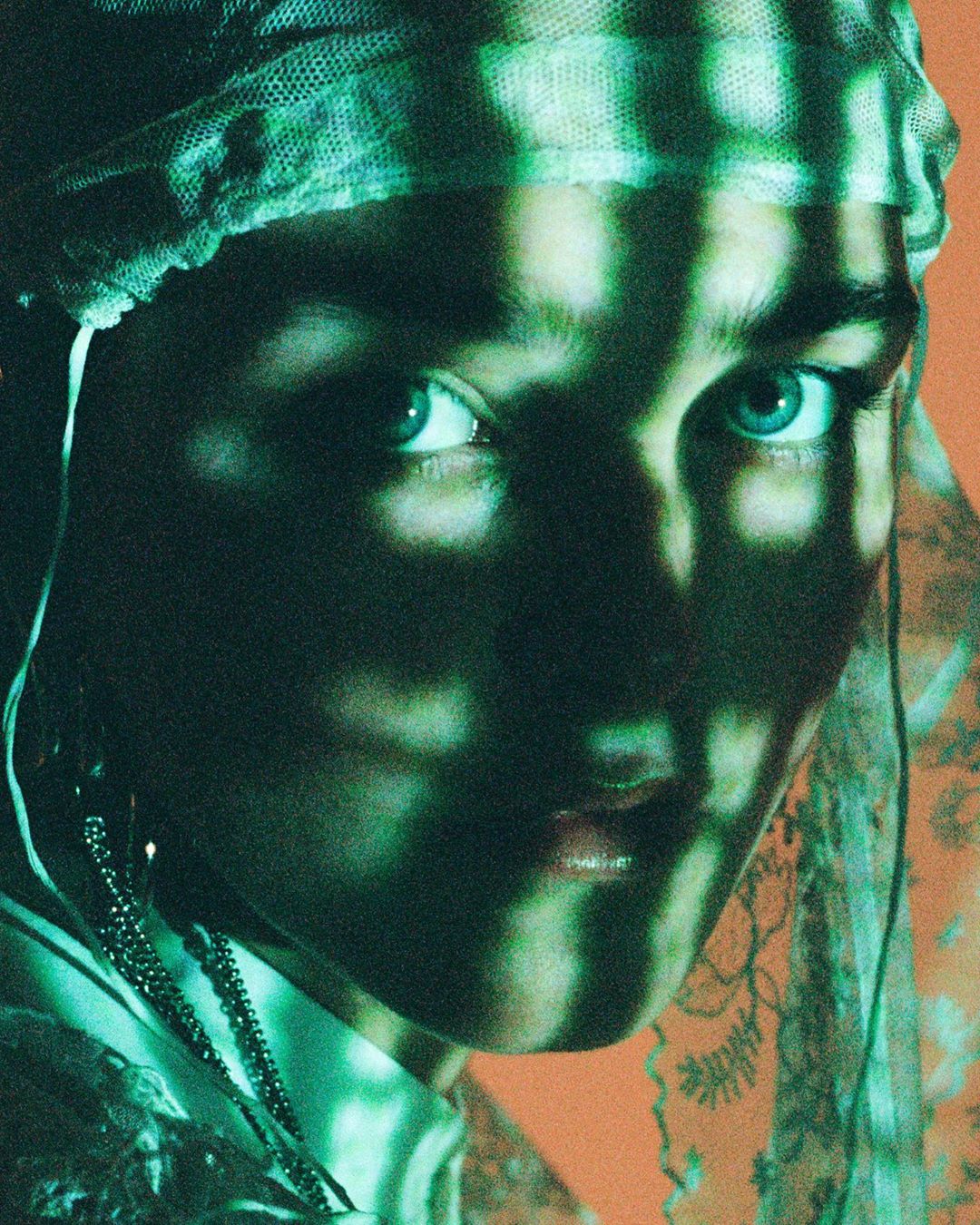 Close-up portrait of a young woman with light skin and blue eyes wearing a patterned headscarf, her face partially obscured by shadow.