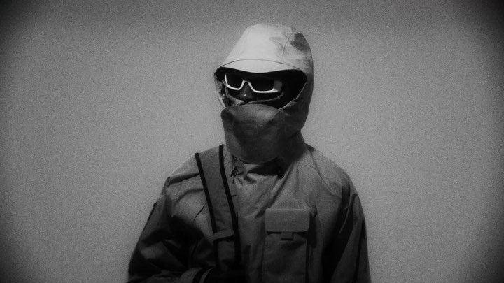 Monochrome portrait of a person wearing futuristic sunglasses, a hooded jacket, and a face covering against a simple background.