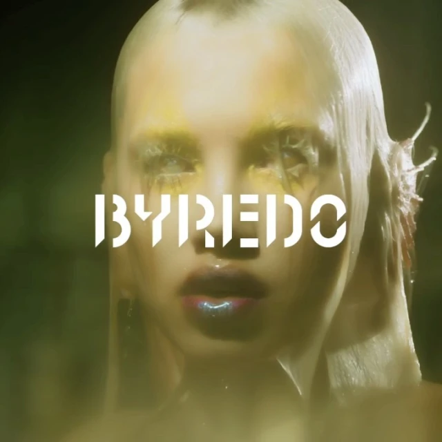 Close-up of a model with dramatic yellow eye makeup, wet-look hair, and glossy lips against a dark green background featuring the BYREDO logo.
