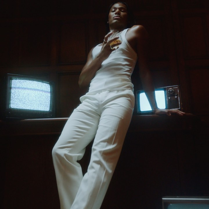 An adult male in a white tank top and pants stands next to vintage television sets, holding a glass, in a dark indoor environment.