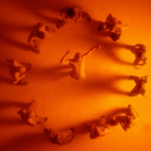 Overhead shot of nine nude figurines positioned in a circle with one in the center, bathed in a warm, diffused orange light.