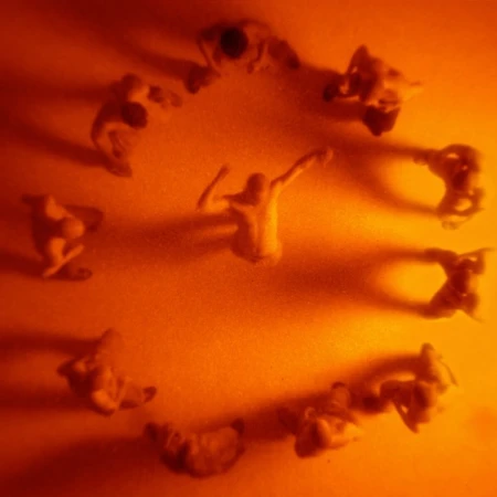 Overhead shot of nine nude figurines positioned in a circle with one in the center, bathed in a warm, diffused orange light.