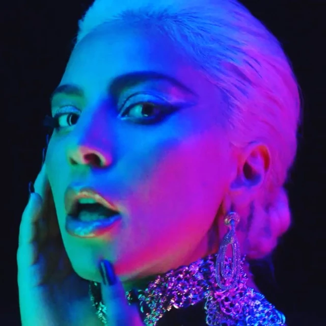 Close-up of an adult female with light blonde hair, dramatic eye makeup, large earrings, and a jeweled collar necklace against a dark background.