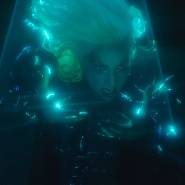 Woman with light blonde hair, expressive face, dark clothing, and bright blue lighting, creating a striking visual composition.