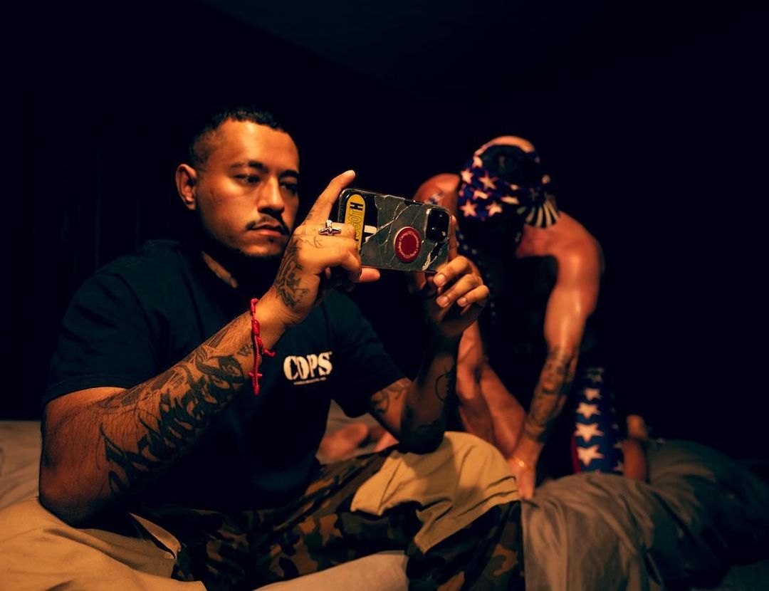A man with arm tattoos holds a phone taking a picture, focusing on another man wearing an American flag bandana over his head. 