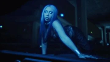 Adult female with blue hair and pale complexion on hands and knees, wearing dark clothing in dim interior environment.