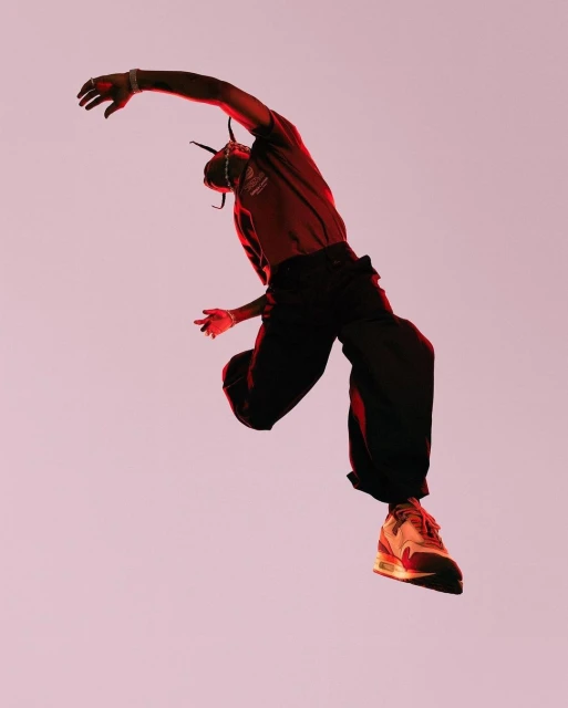 An adult male leaps and poses in mid-air wearing dark trousers, a red t-shirt, and sneakers against a pale pink backdrop.