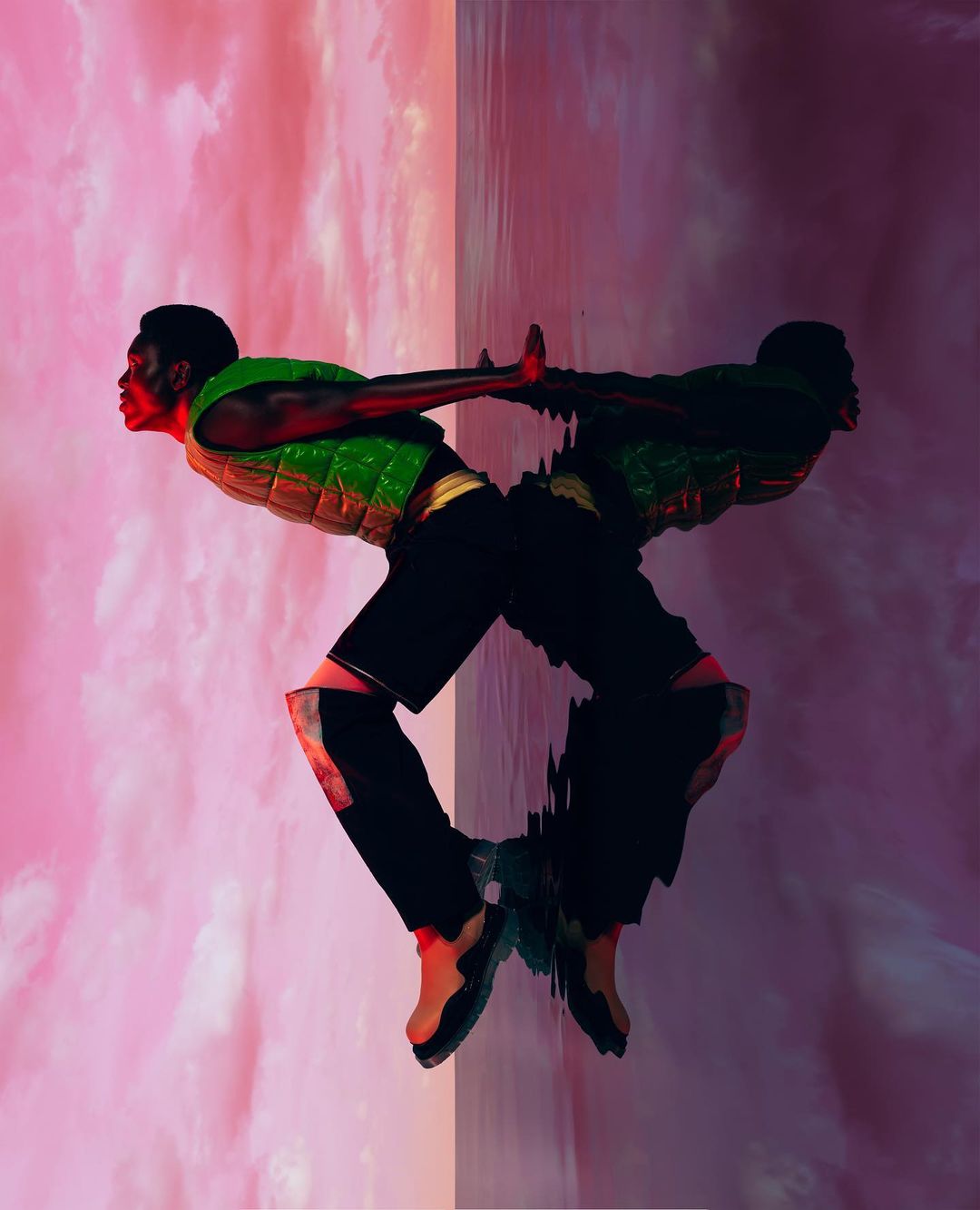 Athletic African American man balances with hand against wall and its reflection; wearing green vest, black shorts, and red-toned athletic shoes.