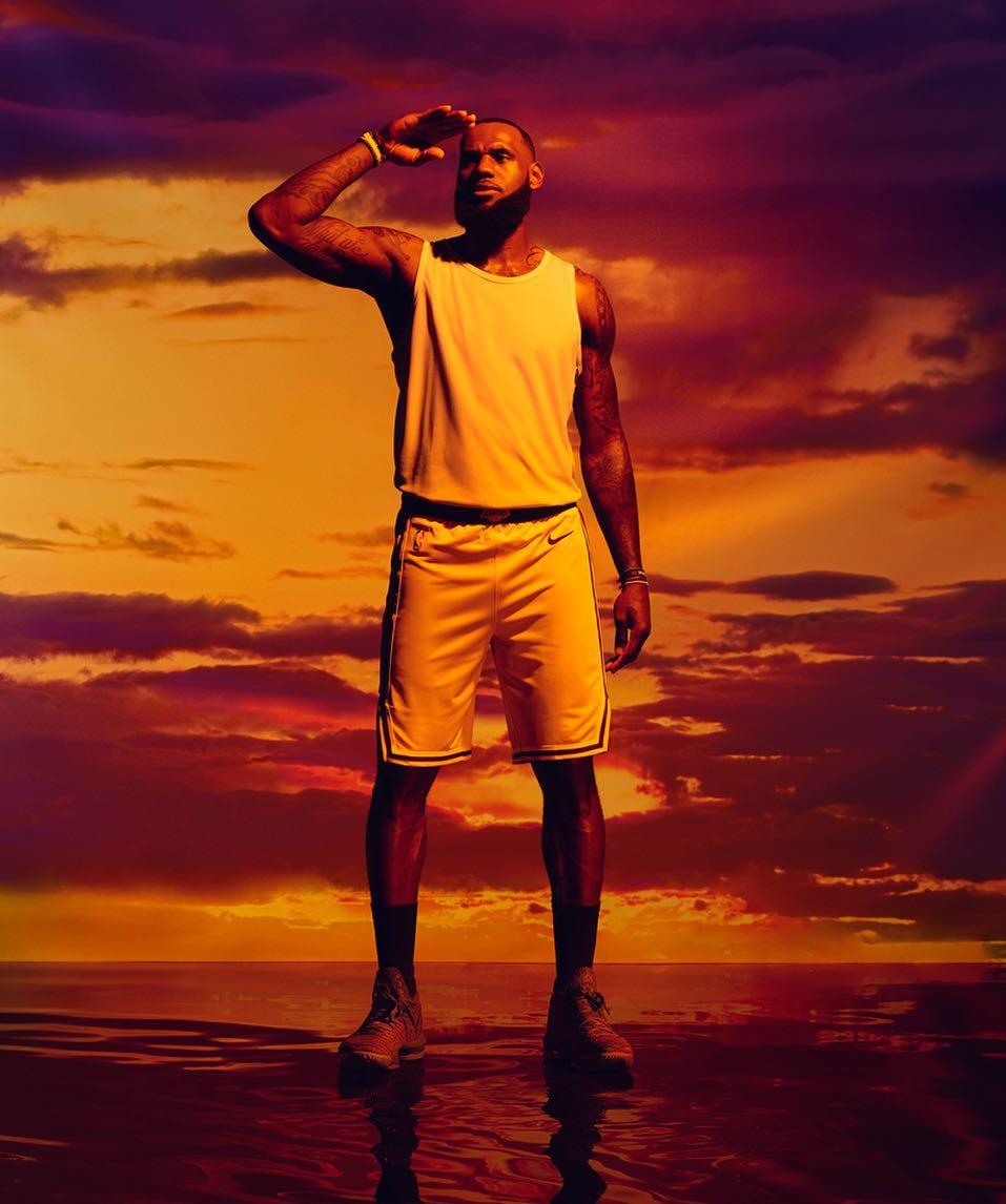LeBron James posing in athletic wear on a water surface against a vibrant sunset sky, looking away and shielding his eyes.