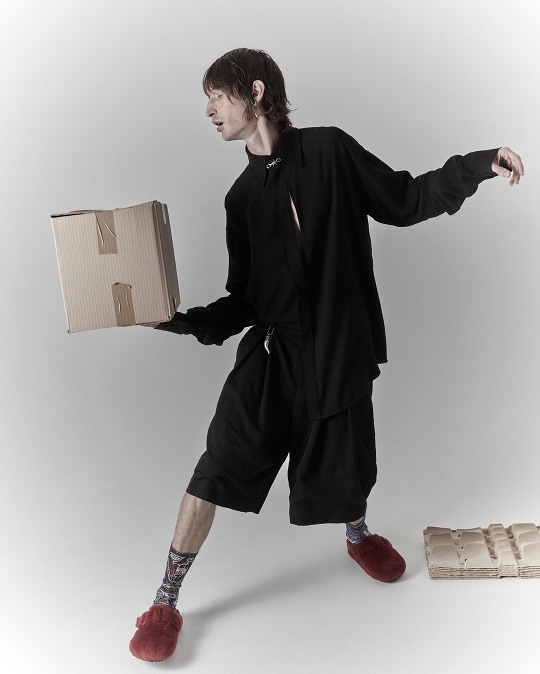Full-length shot of an adult male in a black outfit, holding a cardboard box, wearing red fuzzy slippers, and standing in a bright photo studio.