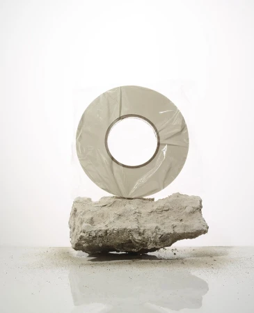 Roll of grey tape resting on a piece of light grey rock against a bright white background in a studio setting with neutral lighting.