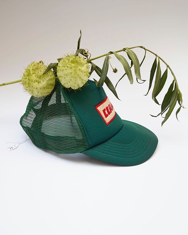 Green trucker hat with green netting back and a plant sprig with two seed pods resting on top. Neutral background.