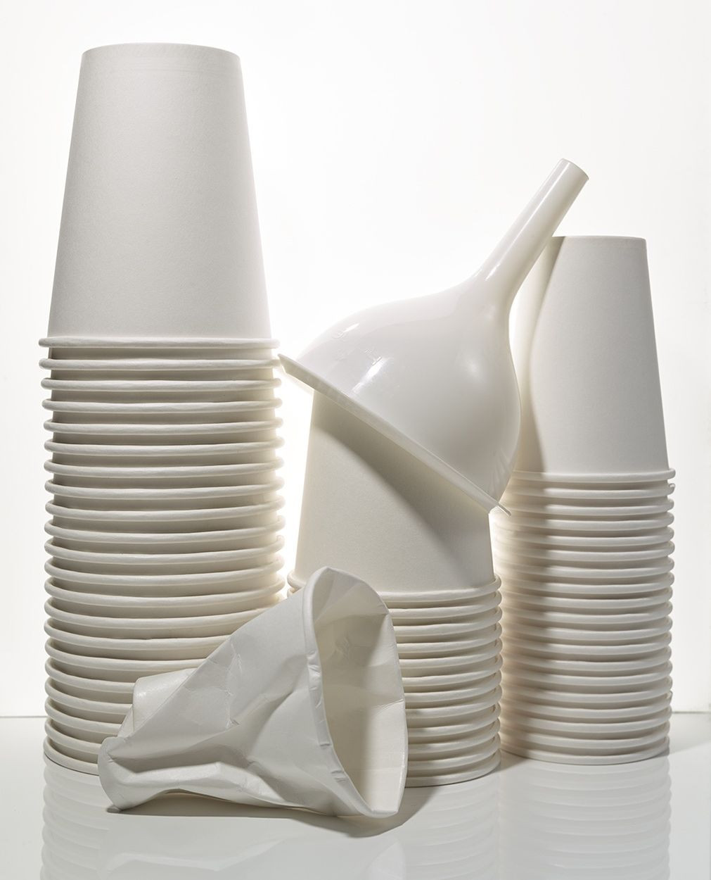 Stacks of white paper cups with a white funnel, one crushed cup in the foreground against a white background, studio lighting.