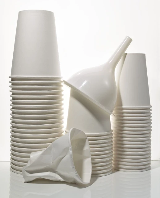Stacks of white paper cups with a white funnel, one crushed cup in the foreground against a white background, studio lighting.