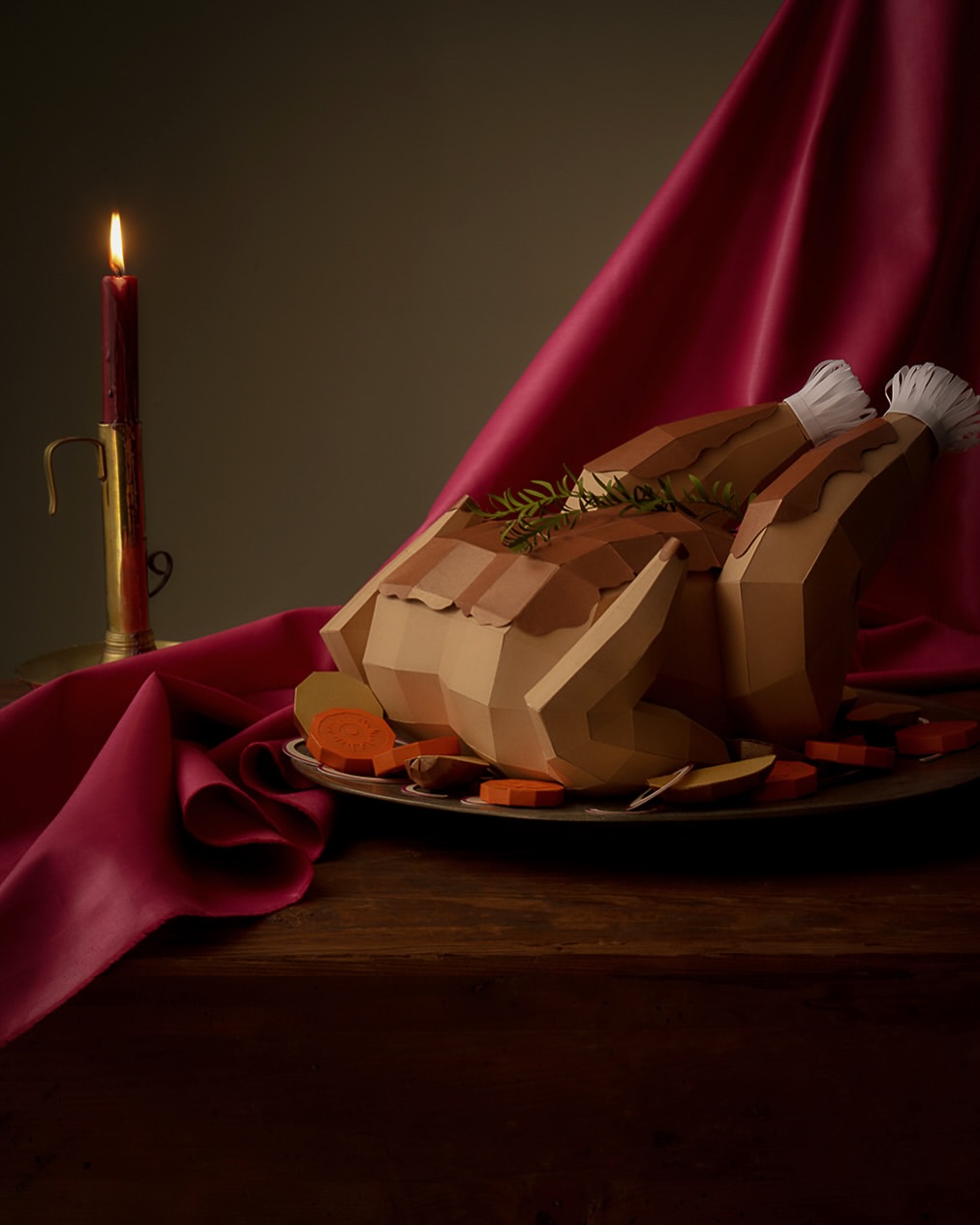 Still life of a crafted paper turkey, carved vegetables, and a lit candle against a draped fabric backdrop.