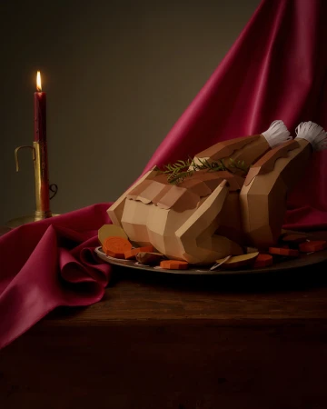 Still life of a crafted paper turkey, carved vegetables, and a lit candle against a draped fabric backdrop.
