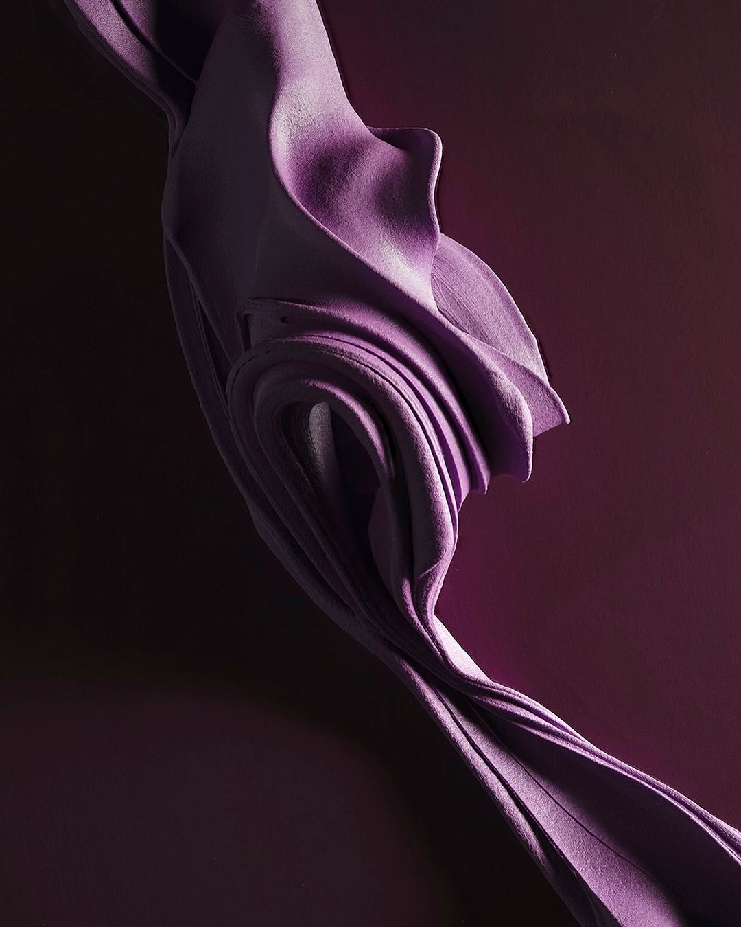 Abstract, flowing fabric in lavender against a dark purple backdrop, showcasing depth and textured textile design in a vertical composition.