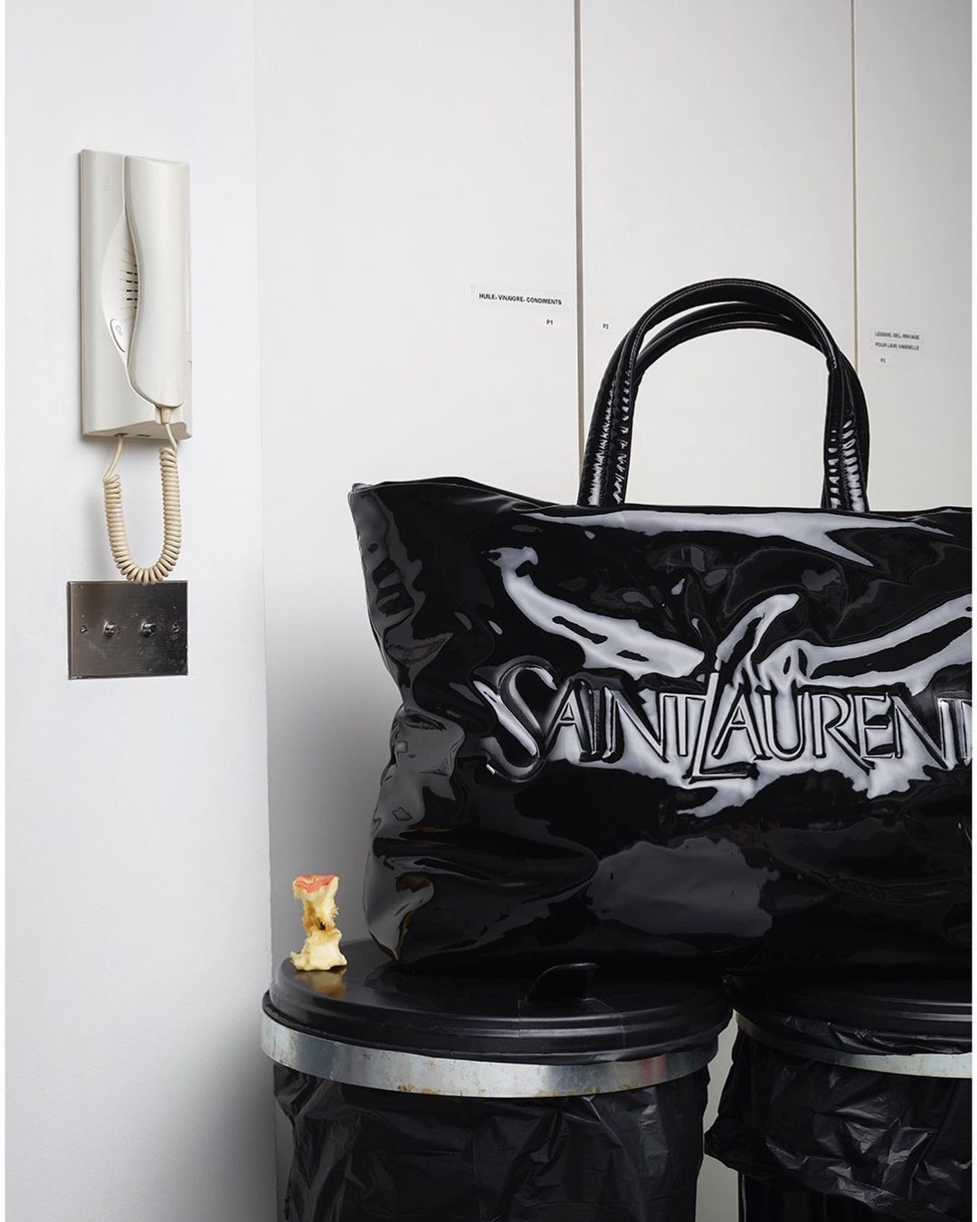 Black Saint Laurent tote bag sits on top of two black trash cans indoors with a white wall, intercomm system, and apple core.