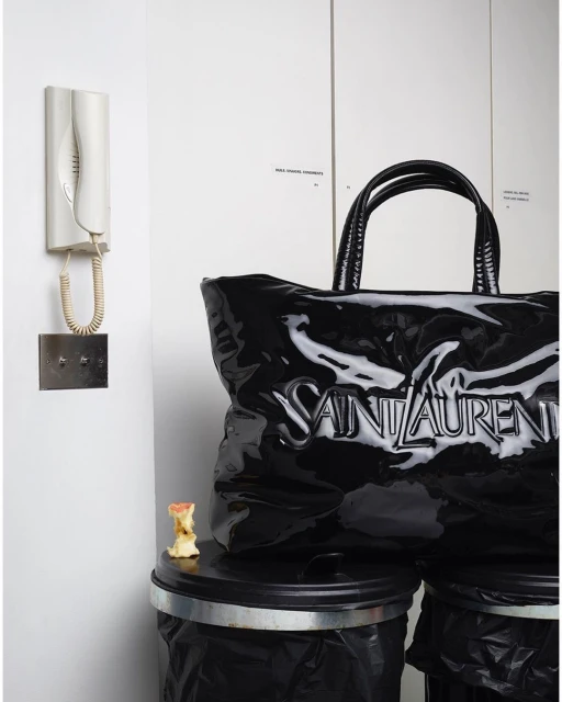 Black Saint Laurent tote bag sits on top of two black trash cans indoors with a white wall, intercomm system, and apple core.