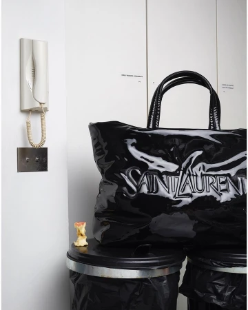 Black Saint Laurent tote bag sits on top of two black trash cans indoors with a white wall, intercomm system, and apple core.