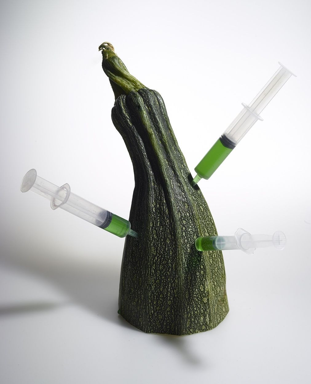 Close-up still life of green zucchini squash with three hypodermic needles injecting it with green liquid, shot in a photo studio.